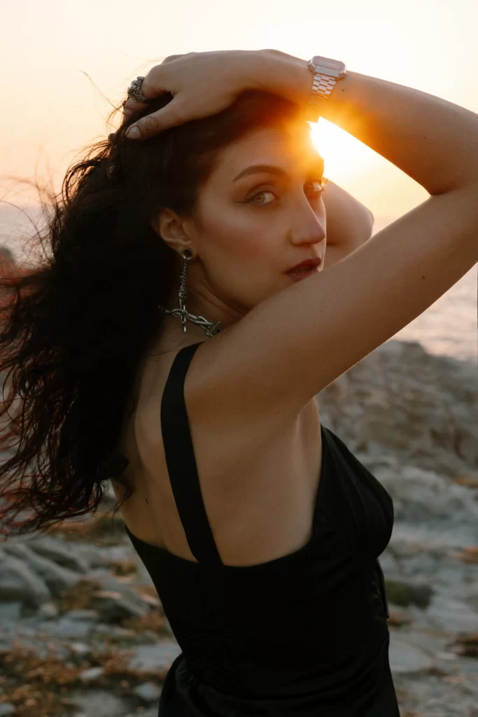 Scenic Photoshoot on Paros Island: A woman striking a picturesque pose on the rocky shores of a Paros beach. Captured by Athens and Paros local photographer Pranvera Dautllari, this Paros photography masterpiece exudes natural beauty