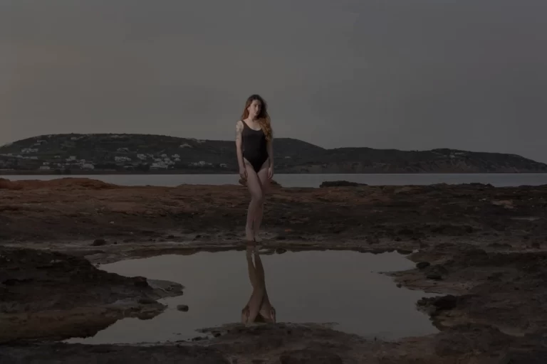 Captivating Paros Island Photoshoot: A woman strikes a stunning pose on the rocky shores of a Paros beach, beautifully captured by Athens and Paros local photographer, Pranvera Dautllari