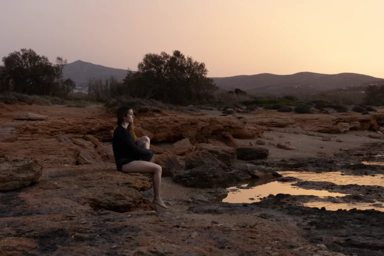 Scenic Paros Island Photoshoot: A woman striking a captivating pose on the rocky shoreline of a Paros beach, captured by Athens and Paros local photographer, Pranvera Dautllari.